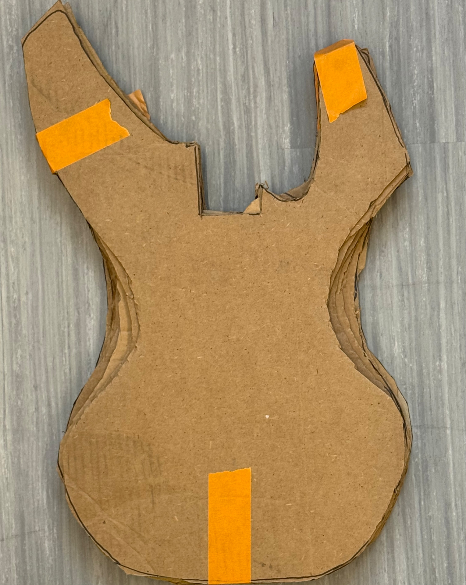 A stack of layered cardboard cutouts of the guitar body, taped together with orange tape on a gray floor