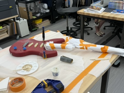 The red 3D-printed guitar body on a paper-lined workbench with its neck wrapped in white paper and orange tape, a bar clamp across the neck joint, and mixed epoxy in a palette with stir sticks nearby