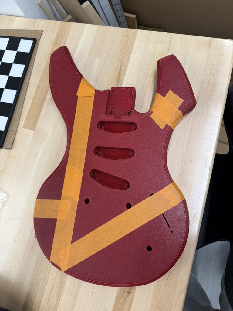 Unfinished 3D-printed electric guitar body in dark red with orange painter's tape strips across it, lying on a wooden workbench