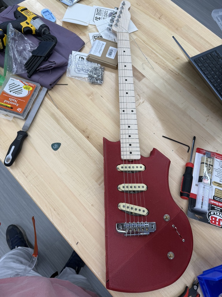 The partially assembled guitar on a workbench with the maple neck bolted on, three cream pickups and a chrome bridge with strings, surrounded by tools and hardware