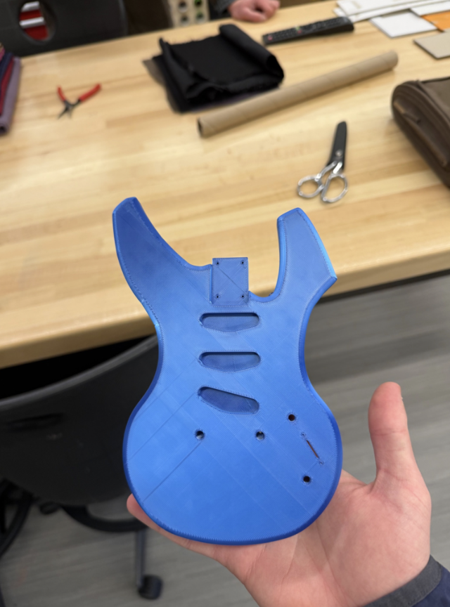 Holding a small blue 1/5 size 3D printed model of the electric guitar body in a workshop